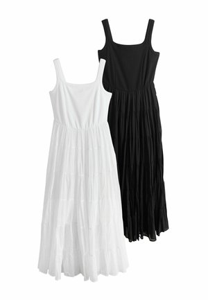 Vrouw met lang donker haar die loopt, gekleed in een mouwloze witte maxi-jurk met lagen en zwarte teenslippers, tegen een effen achtergrond.