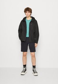 Zwarte lichtgewicht jas met capuchon, turquoise t-shirt, marineblauwe shorts, zwarte sneakers met witte zolen, gecombineerd met zwarte sokken.