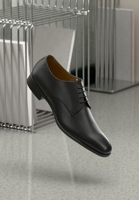 Chaussure formelle en cuir noir suspendue dans les airs à côté d'un présentoir à magazines en métal vertical contenant des livres sur un sol gris.