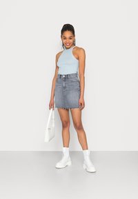 Top sem mangas azul claro, saia mini de denim cinza com bainha desfiada e botas de tornozelo brancas com cordões. Segurando uma mala branca.