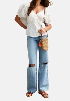 White puff-sleeve blouse with eyelet detailing, paired with light blue flared jeans featuring distressed knees and brown slide sandals. Woven bag included.