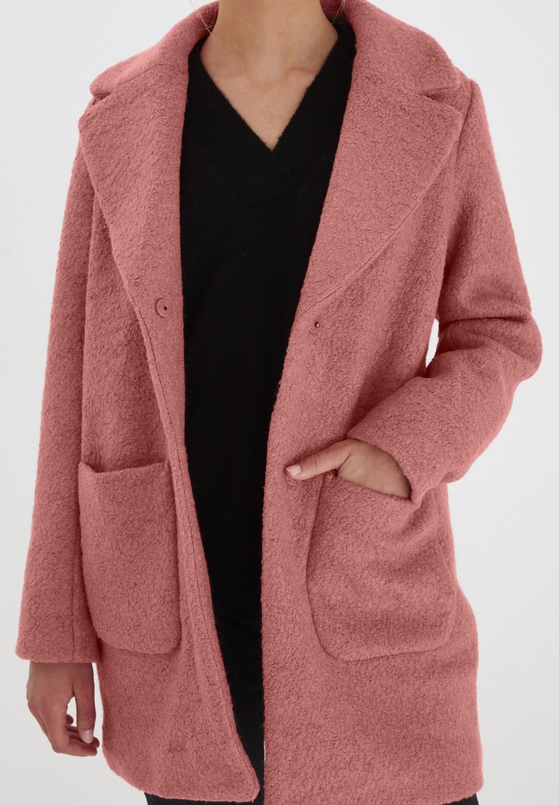 Femme portant un manteau texturé rose poussiéreux avec de grandes poches avant par-dessus un haut noir à col en V, main dans la poche droite, devant un fond uni.