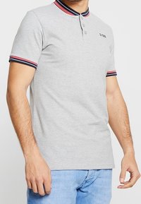 Camisa polo gris de algodón con mangas cortas, que presenta un cuello con franjas rojas y marinas, dos botones y un pequeño logo en el pecho.