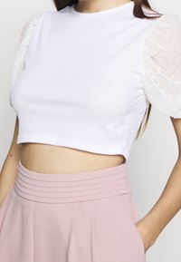 Bílý žebrovaný crop top s průhlednými bílými balónovými rukávy s puntíky, spárovaný s vysokými světle růžovými kalhotami s sklady.