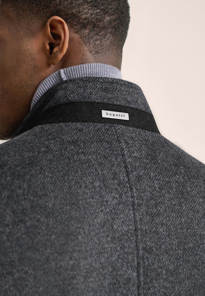 Cappotto grigio testurizzato con un'etichetta nera che presenta il nome del marchio "bugatti" sul colletto. Collo interno a maglia visibile, che enfatizza il design a strati.