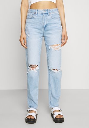 Mujer con vaqueros desgarrados azul claro y sandalias blancas, de pie frente a un fondo claro y sencillo, torso visible desde la cintura hacia abajo.