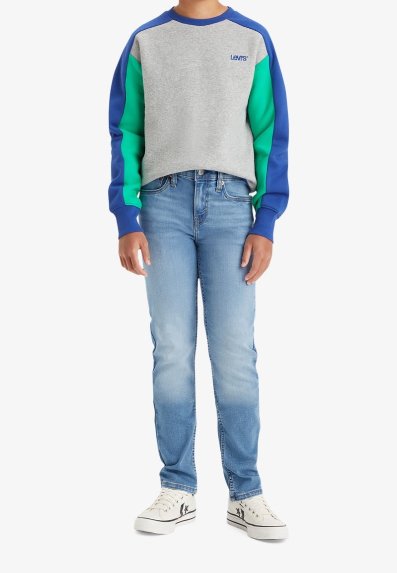 Personne portant un sweat Levi's bleu-vert-gris, un jean bleu clair et des baskets blanches avec des détails étoiles noires, debout devant un fond blanc.