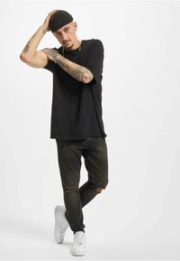 Melnā T-kreklā, nolietotos tumšajos džinsos, baltās sneakeres un melnā adītā cepure. Redzami tetovējumi uz rokām, modele pozē ar vienu roku uz galvas.