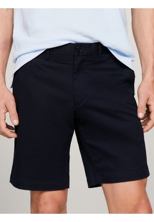 Shorts noirs pour hommes, taillés sur mesure, avec fermeture par bouton et zip, portés avec une chemise bleu clair rentrée à la taille.
