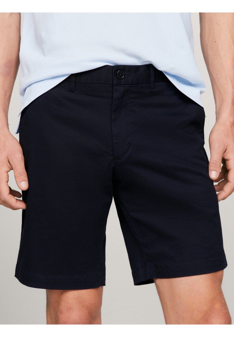 Shorts noirs pour hommes, taillés sur mesure, avec fermeture par bouton et zip, portés avec une chemise bleu clair rentrée à la taille.