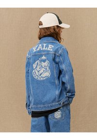 Veste en denim bleu clair avec le texte blanc "YALE" et un graphique de bulldog au dos. Elle possède un col classique et des poches avant.