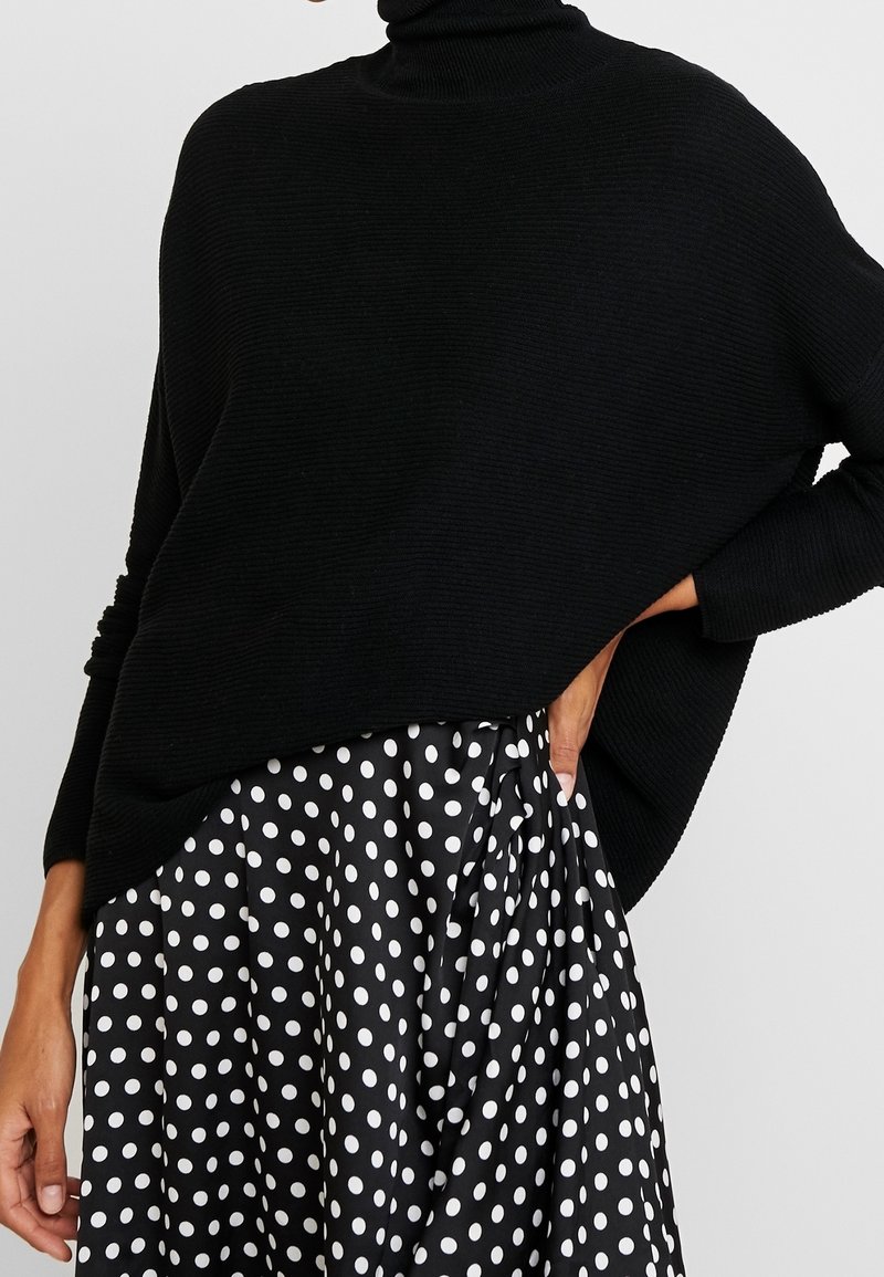 Un pull à col roulé noir à côtes porté par-dessus une jupe à pois noir et blanc. Le pull a une coupe décontractée avec des épaules tombantes et des manches longues.