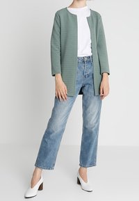 Ljusgrön texturerad kofta över en vit t-shirt, kombinerad med ljusblå jeans med raka ben och vita högklackade skor.