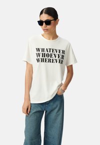 T-shirt en coton blanc avec le texte noir en gras "QUOI QUE CE SOIT, QUI QUE CE SOIT, OÙ QUE CE SOIT." Associé à un pantalon en denim bleu et des lunettes de soleil noires.