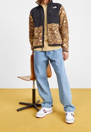 Mężczyzna ubrany w brązowo-czarną wzorzystą kurtkę North Face, beżową koszulę, jasnoniebieskie dżinsy i białe tenisówki, stojący obok drewnianego krzesła.