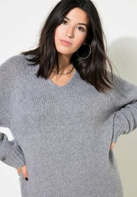 Pull en tricot gris avec un col en V, coupe oversize, poignets côtelés et texture douce. Présente des manches amples et une silhouette décontractée.