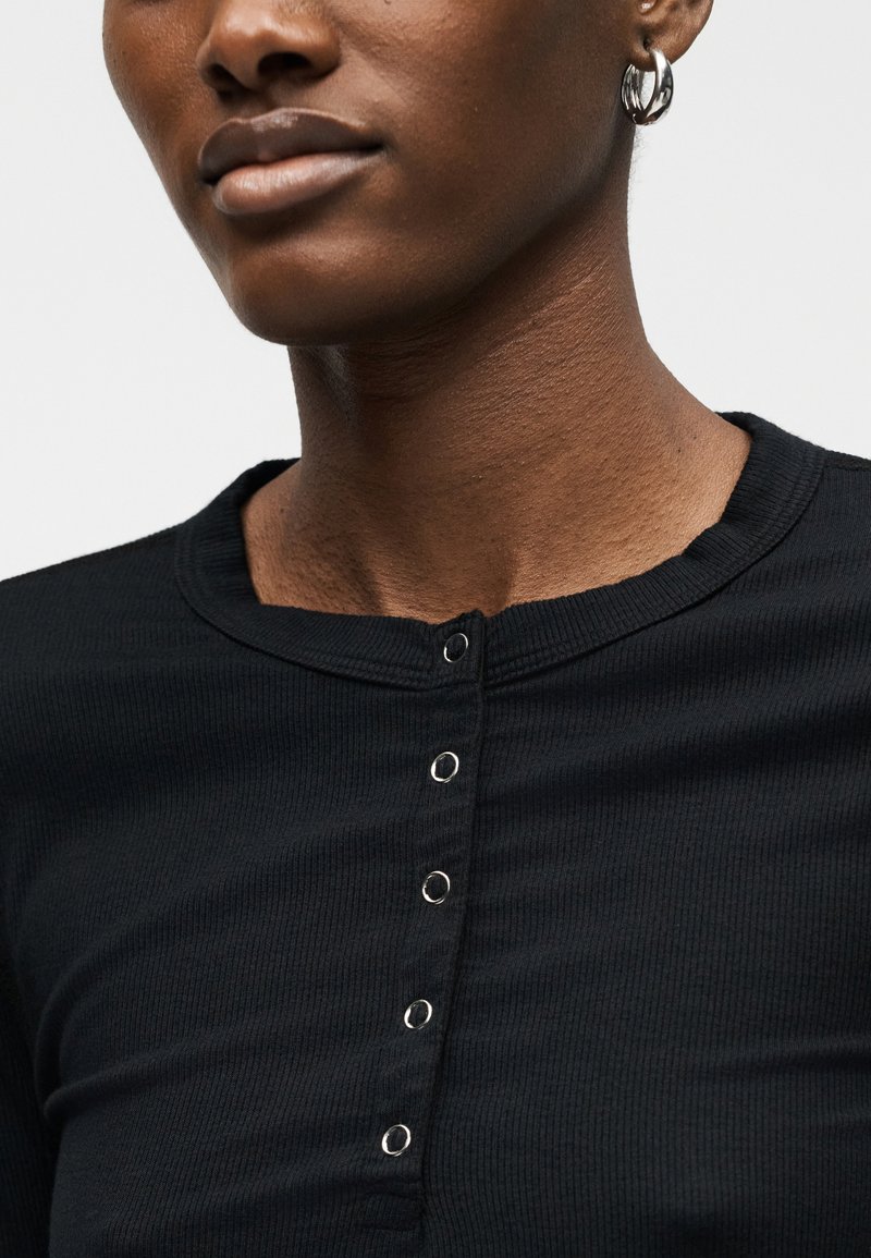 Close-up of a person wearing a black ribbed henley shirt with silver snap buttons and a silver hoop earring.