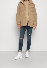 Beige Fleecejacke aus weichem Plüsch mit Knöpfen und zwei Brusttaschen, darüber ein hellbeiger Strickpullover, kombiniert mit dunklen, zerrissenen Jeans und weißen Turnschuhen.
