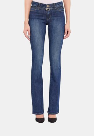 Donna che indossa jeans blu a gamba dritta con passanti per cintura e scarpe nere con tacco alto, in piedi su sfondo bianco.