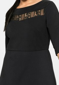 Robe noire avec un panneau en dentelle à l'avant supérieur, des manches trois-quarts, un buste structuré et une texture de tissu lisse.