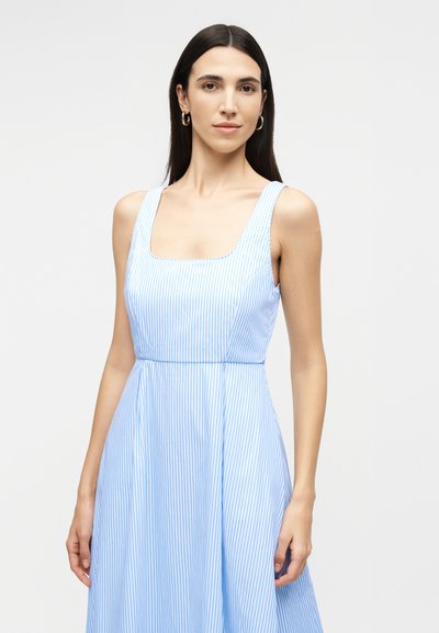 Femme aux longs cheveux foncés portant des boucles d'oreilles créoles en or et une robe sans manches bleu clair à fines rayures verticales, sur un fond uni.