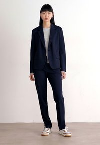 Námornický blazer a kalhoty, kombinované se šedou košilí a bílo-hnědými teniskami. Hladká tkanina, přizpůsobený střih, jednoduché detaily knoflíků.