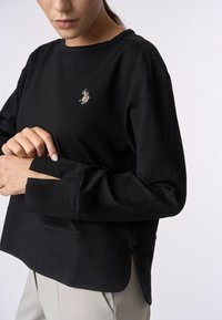 Felpa nera con maniche lunghe, scollatura rotonda, spacchi laterali e un piccolo logo ricamato sul petto. Tessuto morbido.