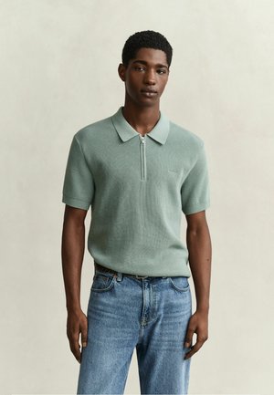 Jeune homme aux cheveux courts et bouclés porte un polo zippé vert clair à manches courtes et un jean bleu, sur un fond clair et uni.