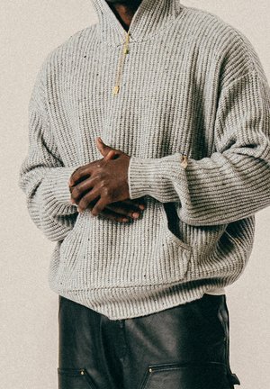 Homme portant un sweat à capuche en maille grise et un pantalon noir, les mains jointes à la taille devant un fond neutre.
