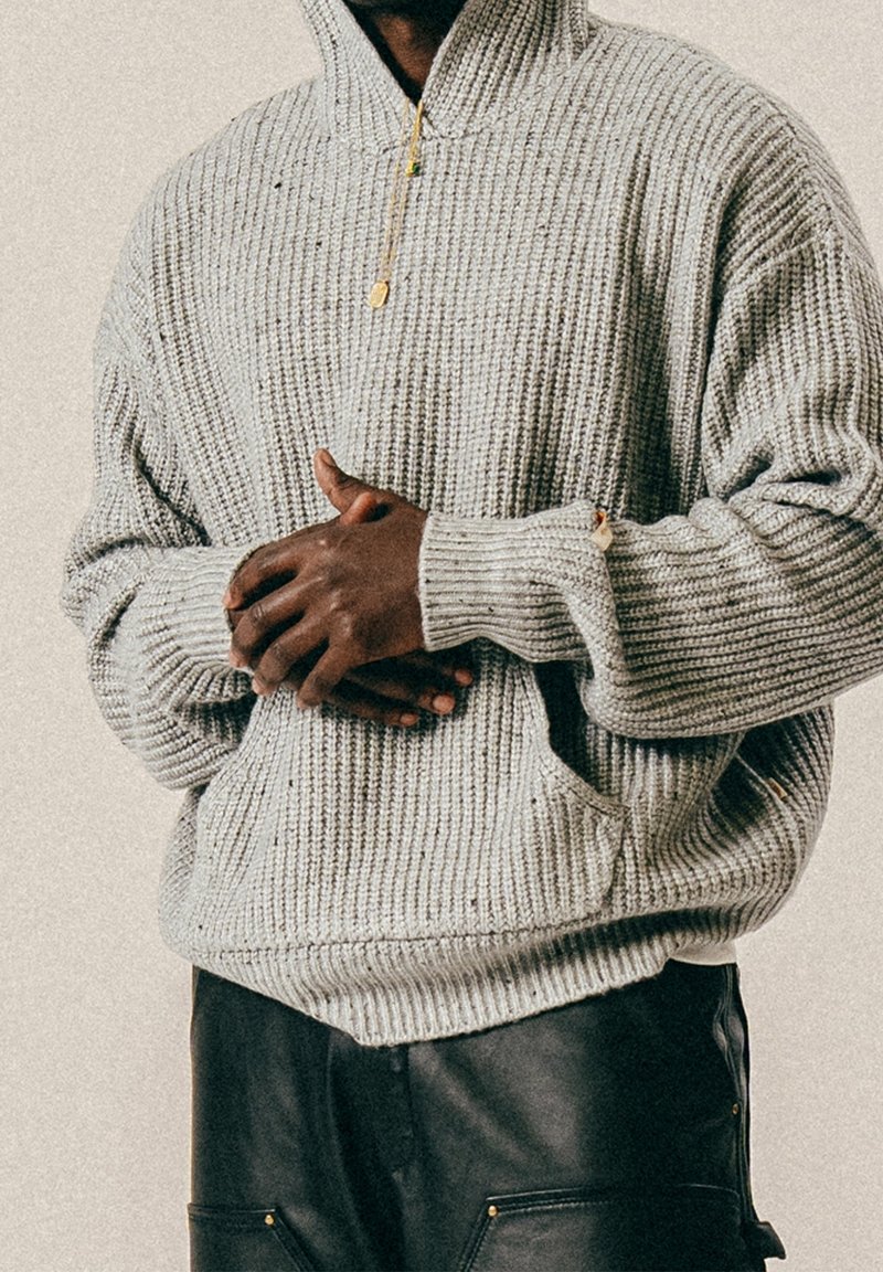 Homme portant un sweat à capuche en maille grise et un pantalon noir, les mains jointes à la taille devant un fond neutre.