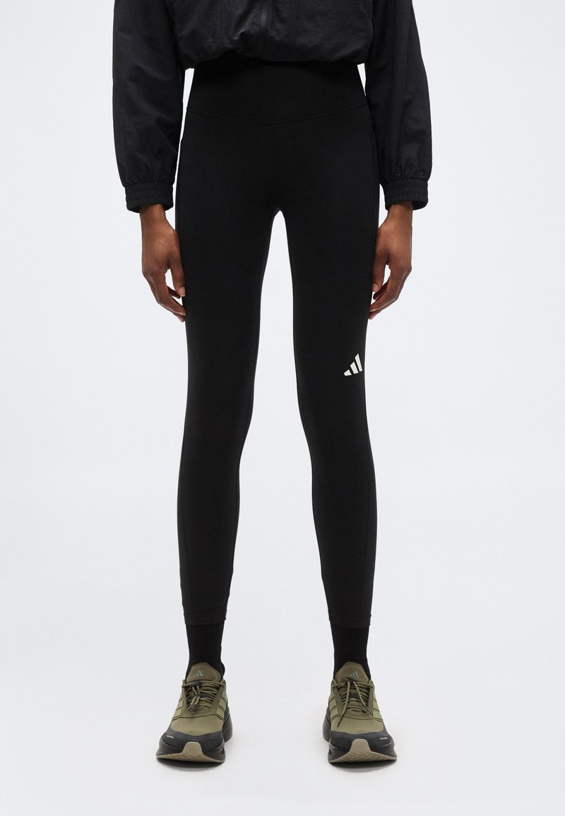 Άτομο που φοράει μαύρα κολάν ψηλής μέσης με λευκό λογότυπο στη δεξιά μηρό και ελιά πράσινα αθλητικά παπούτσια, στέκεται μπροστά σε απλό φόντο.