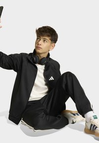 Teenager in schwarzem Adidas-Trainingsanzug und Kopfhörern, der ein Selfie macht, während er auf dem Boden sitzt.