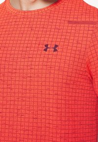 Primer plano de una persona vistiendo una camiseta deportiva roja con un patrón y un logo negro de Under Armour centrado en el pecho.