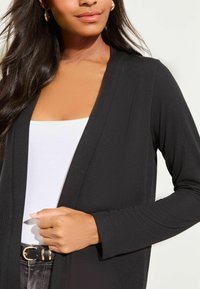 Cardigan noir à manches longues en tissu doux, design ouvert à l'avant, porté sur un débardeur blanc, avec une ceinture noire visible à la taille.