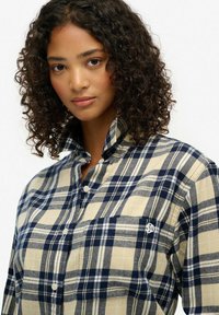 Chemise à carreaux beige et marine avec un design à boutons, un col montant et une poche avant. Présente un logo discret sur la poche.