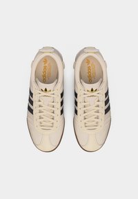 Beiget väriset Adidas-tennarit mustilla raidoilla, kultaisella logolla kielen päällä ja nauhoilla, kuvattuna ylhäältä valkoisella taustalla.