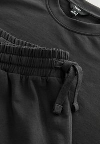 Tricou din bumbac negru și pantaloni de training cu talie elastică. Caracteristici detaliu cu șnur și textură netedă. Design simplu, ambele în culoare închisă.