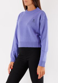 Maglione corto viola con scollo a coste e maniche con polsini; presenta un piccolo logo nero ricamato sul petto. Indossato con pantaloni neri.