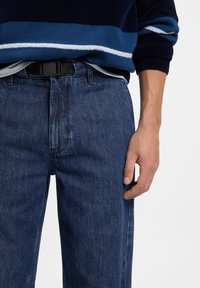 Pull en tricot bleu avec des rayures blanches et bleu clair, associé à un jean denim bleu foncé, avec une ceinture noire, gros plan sur la taille et la main.