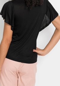 Woman wearing a black short-sleeve top with flutter sleeves and light pink pants, hand resting on waist, shown from back.