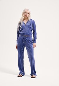 Kék velúr szabadidőruha, cipzáras, rövidített kapucnis pulóverrel és hozzáillő nadrággal, oldalsó csíkkal és gumírozott derékrésszel.