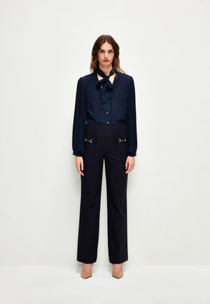 Marinebluse mit einer Bow-Tie-Kragen, langen gefalteten Ärmeln und Knöpfen; kombiniert mit hoch taillierten Marinehosen mit vorderen Taschen und silbernen Beschlägen.