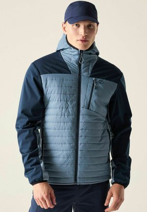 Young man wearing a blue and gray quilted jacket with a zip-up hood and a navy blue cap, standing against a plain light background.