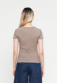 Femme avec de longs cheveux ombrés portant un haut beige en rib de courte manche ajusté et un jean bleu foncé ample, vue de dos contre un fond uni.