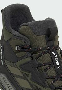 Schwarzer und olivgrüner Wanderschuh aus strukturiertem Stoff, mit gepolsterter Lasche, kontrastierenden orangefarbenen Nähten und Gummielementen. Verfügt über ein Schnürdesign.