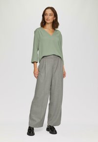 Blouse verte légère avec un décolleté en V et des plis aux épaules, associée à un pantalon ample en chevron gris et des chaussures à plateforme noires.