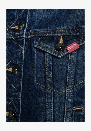 Voorklepzak van een spijkerjasje met knoopsluiting, gele stiksels en een rood stoffen label met de tekst "BIG STAR".