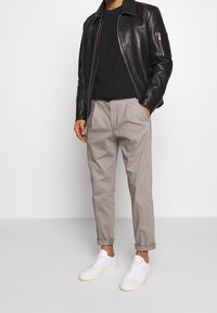 Zwarte leren jas, grijze geplooide broek en witte sneakers. De jas heeft een ritssluiting en een opstaande kraag. Over het geheel genomen een casual pasvorm.