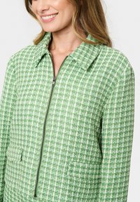 Zelený tvídový kabát se zipem vpředu, límcem a texturovaným vzorem s mřížkovým tkaním. Součástí jsou dvě boční kapsy.