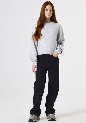 Lichtgrijze gebreide sweater met brede mouwen, gecombineerd met zwarte broek met een subtiel golvend patroon. Grijze sneakers maken de outfit compleet.